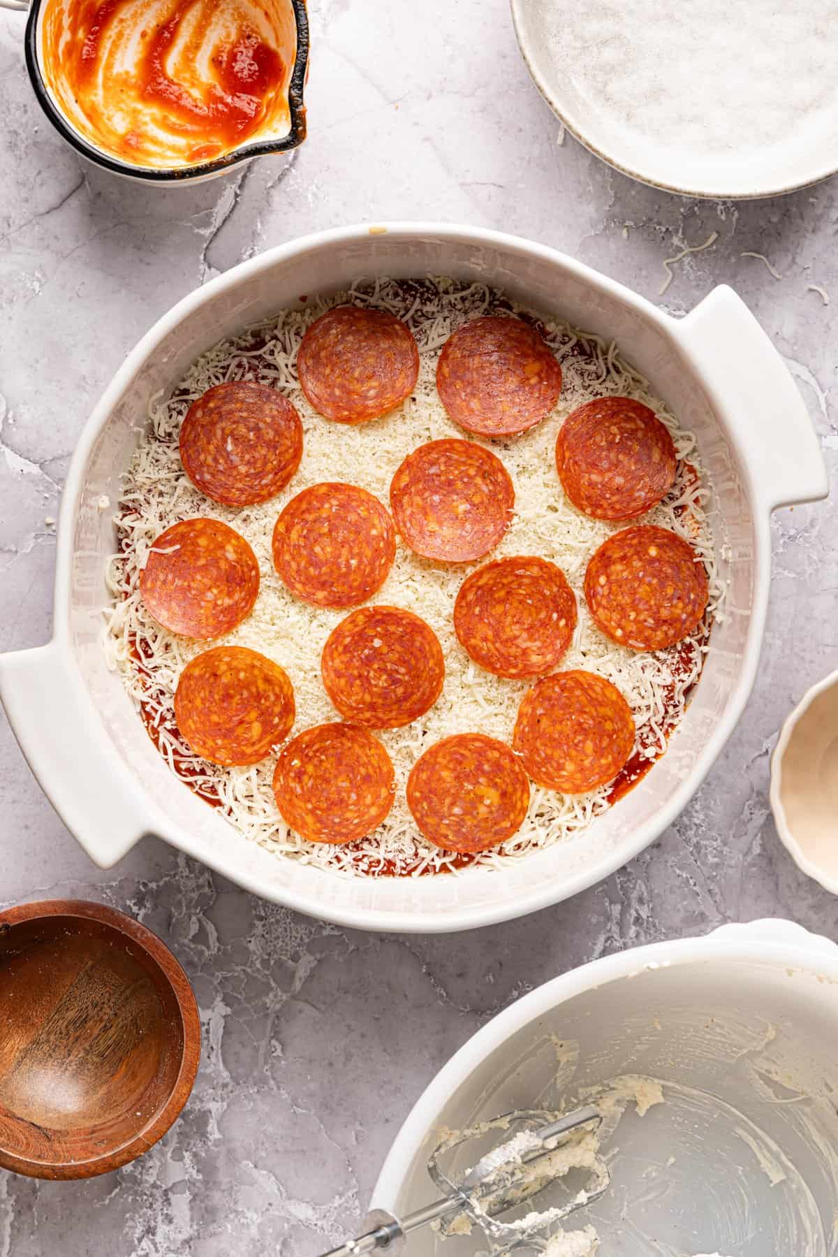 Unbaked high protein pizza dip in a baking dish with shredded cheese and pepperoni layered on top.