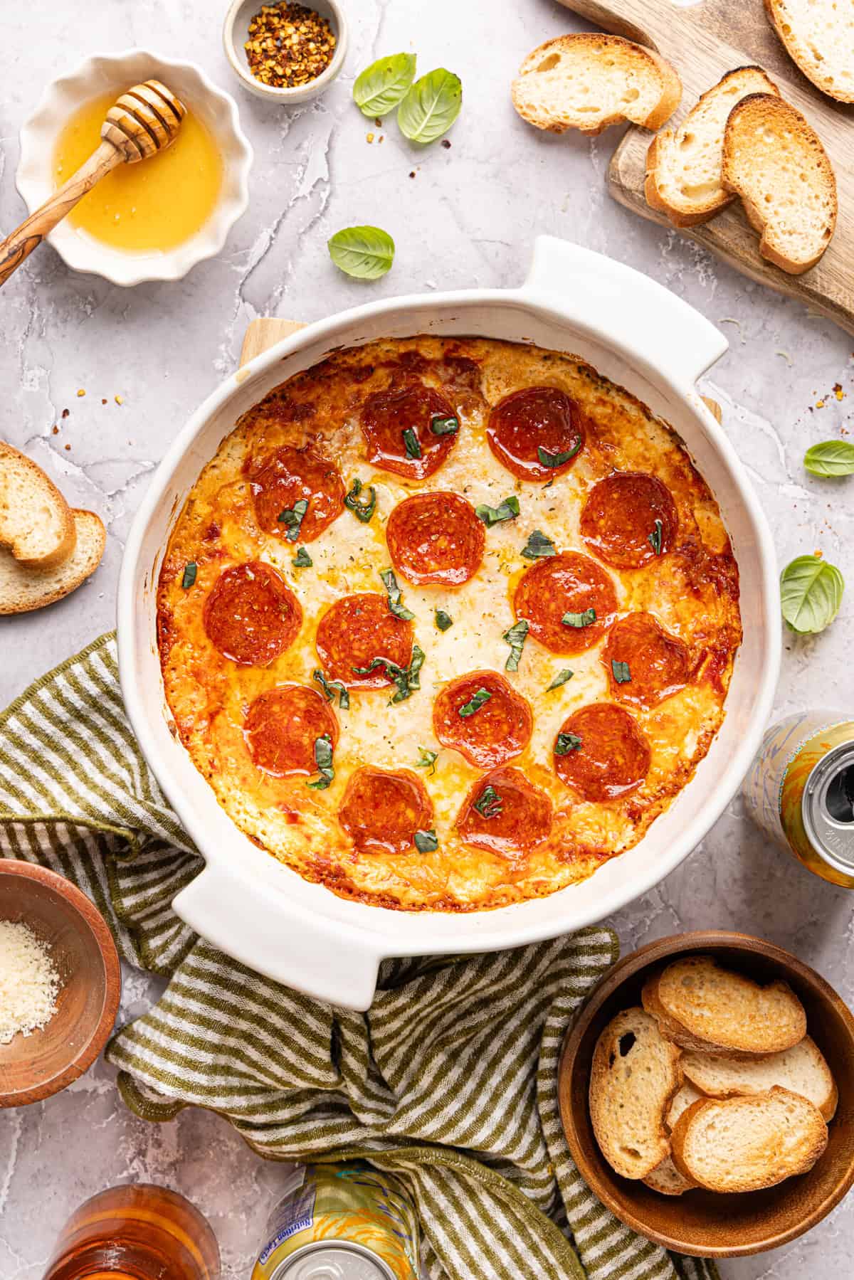 Baked high protein pizza dip in a round white dish, topped with pepperoni, melted cheese, hot honey drizzle, and fresh basil.