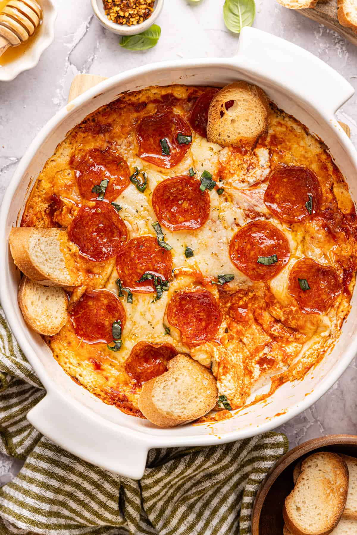 Baked high protein pizza dip in a round white dish, topped with pepperoni, melted cheese, hot honey drizzle, and fresh basil. A few small slices of bread are resting in the dip.