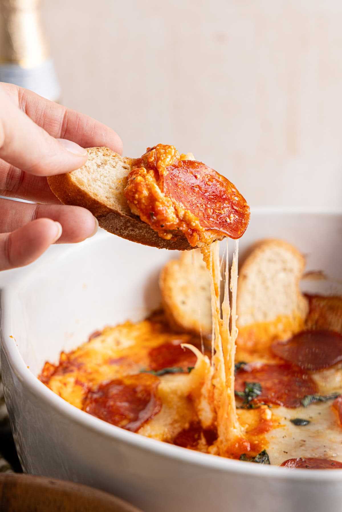 Hand holding a slice of bread topped with a scoop of high protein pizza dip, with the rest of the dip in the dish below.