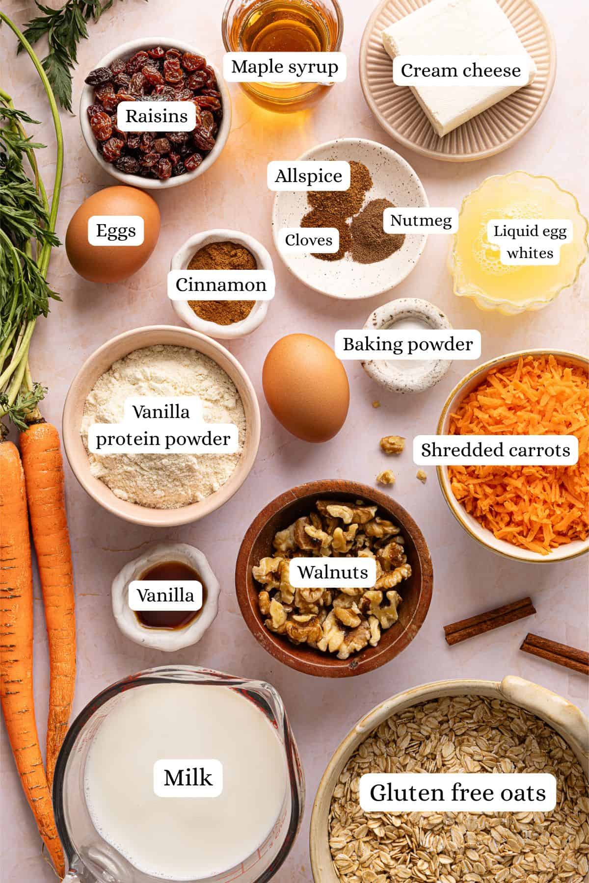 Ingredients for carrot cake baked oats labeled and laid out in nesting bowls on a light pink countertop.