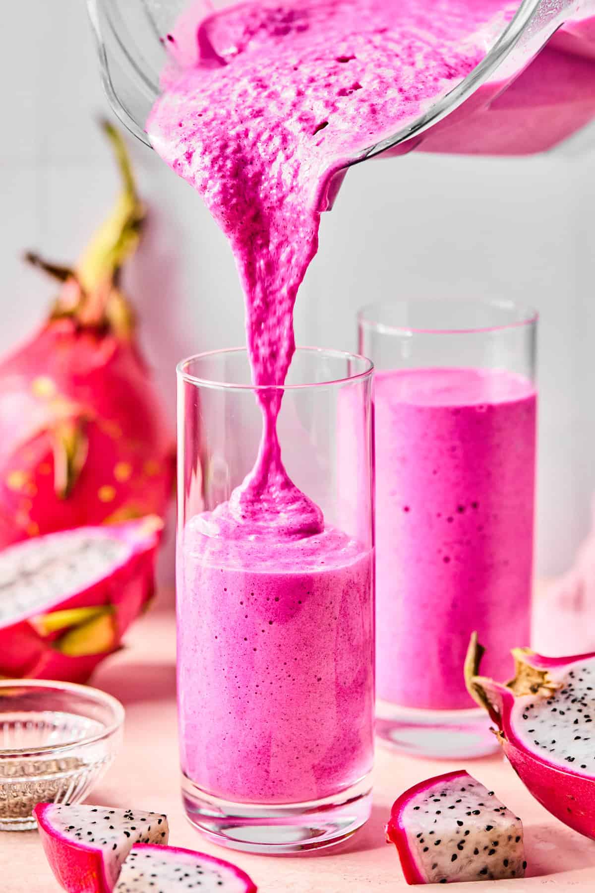 Dragonfruit smoothie being poured into a tall glass with another full glass behind it and sliced dragonfruit scattered around.
