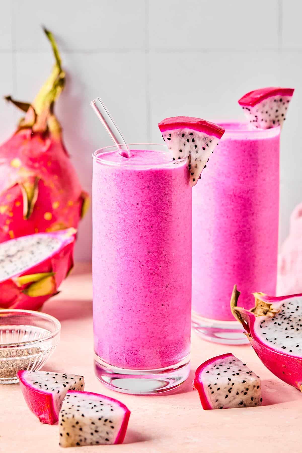 Two tall glasses of dragonfruit smoothie with glass straws and fresh sliced dragonfruit on the side.