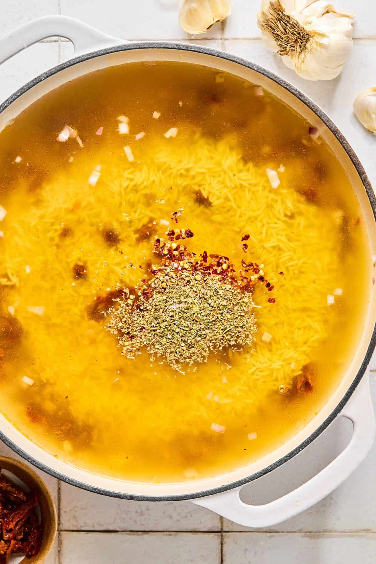 Gluten-free orzo simmering in chicken broth with Italian seasoning and red pepper in a skillet.
