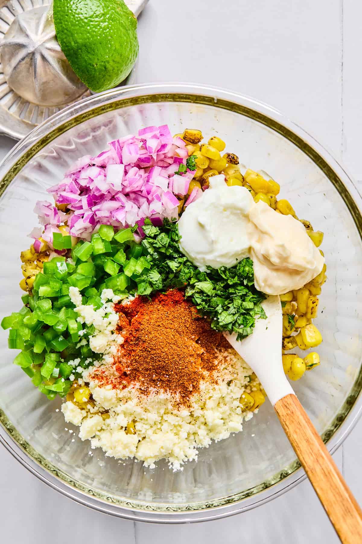 Street corn mixture being stirred with red onion, jalapeño, cilantro, mayo, Greek yogurt, lime juice, spices, and cotija cheese in a glass bowl.