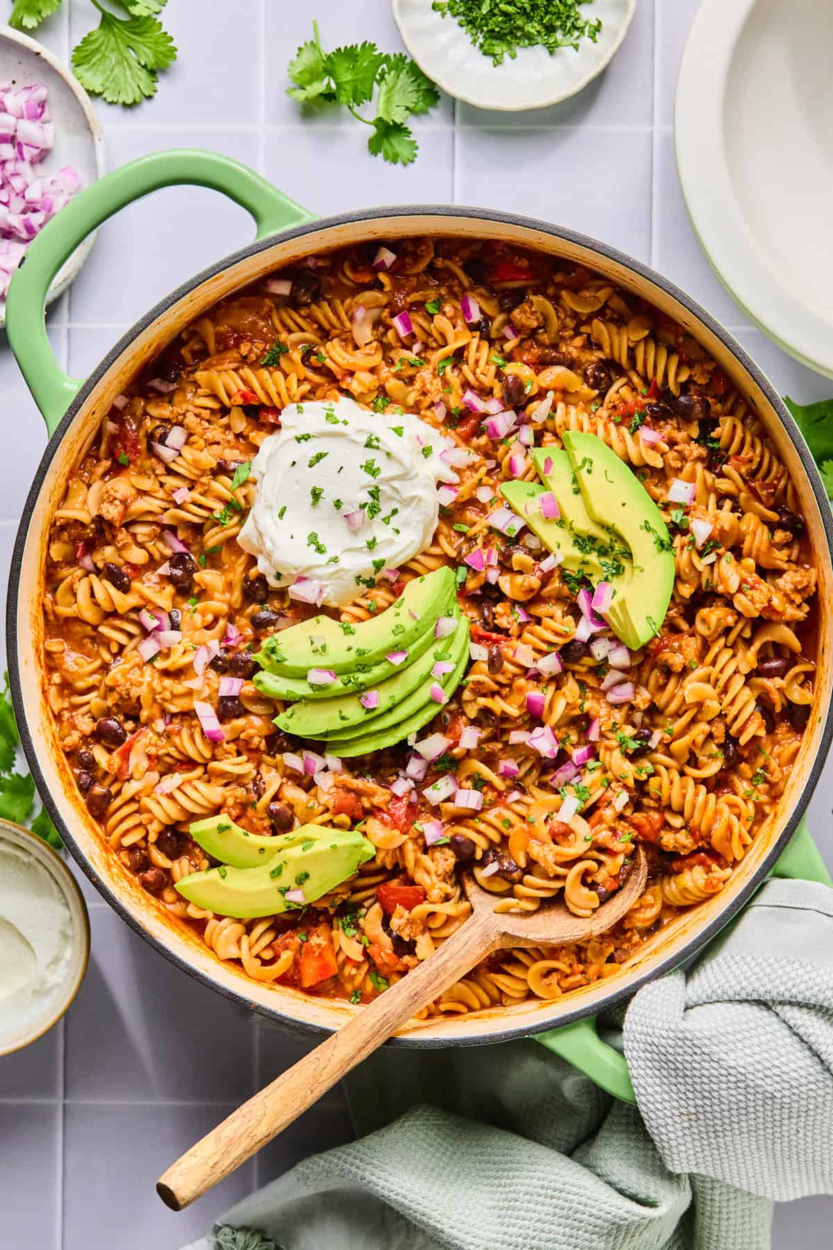 One pot taco pasta in the pot topped with sour cream, avocado, and cilantro with a wooden spoon.
