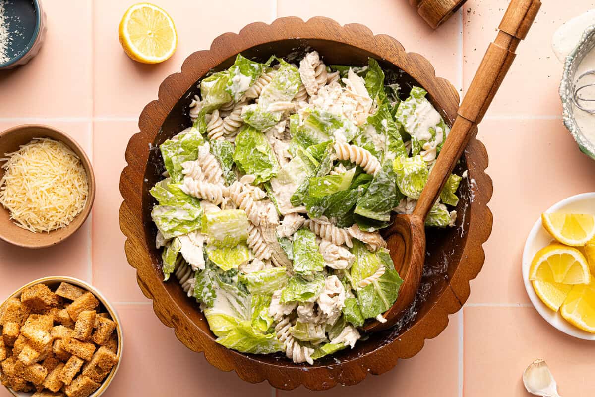 chicken caesar pasta salad tossed together in a wooden bowl with a wooden spoon