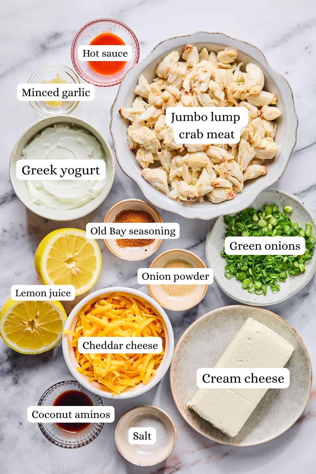 Crab dip recipe ingredients labeled and laid out in nesting bowls on a light gray and white countertop.