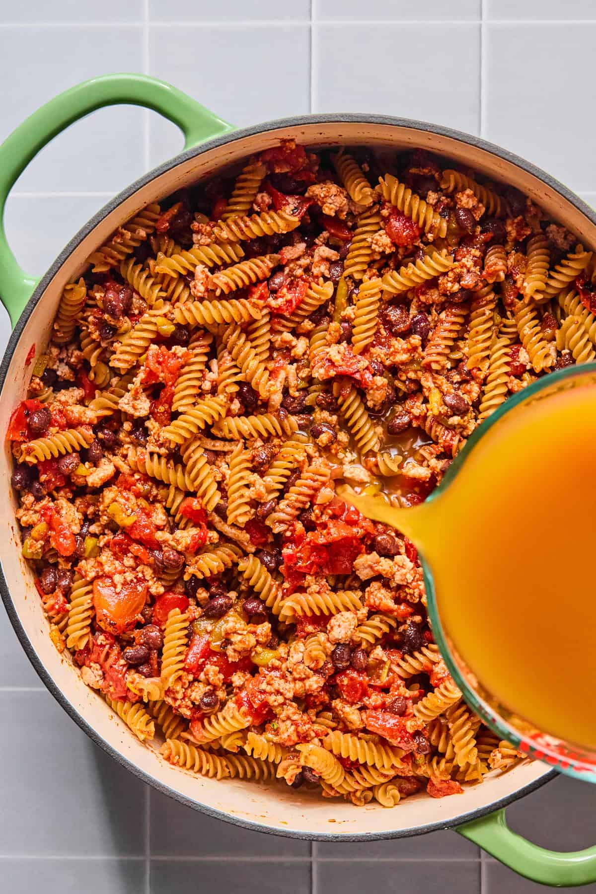 Chicken broth being poured into the pot with the taco seasoning, tomato paste, diced tomatoes, green chiles, black beans, and pasta.