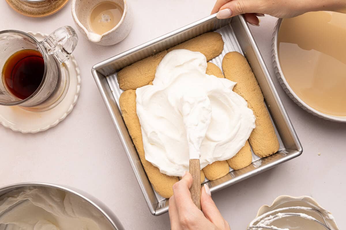whipped cream being spread on lady fingers in a pan