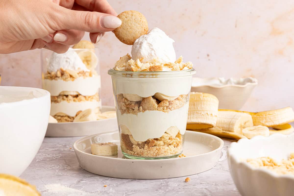 a hand placing a vanilla wafer on top of a cup of banana pudding