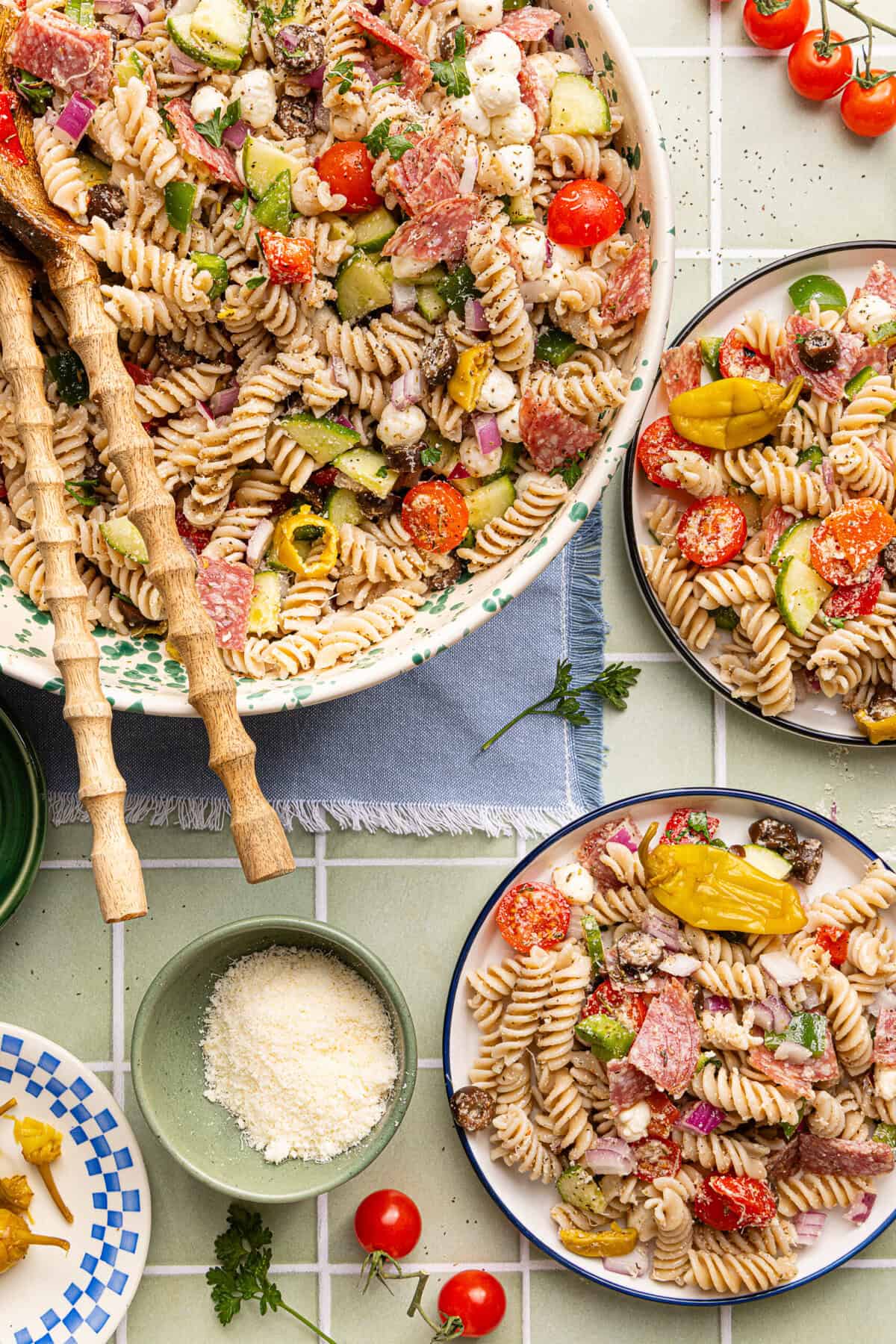 Gluten free pasta salad served in a decorative bowl with wooden serving spoons, alongside two plates of pasta salad and a small bowl of grated parmesan cheese.