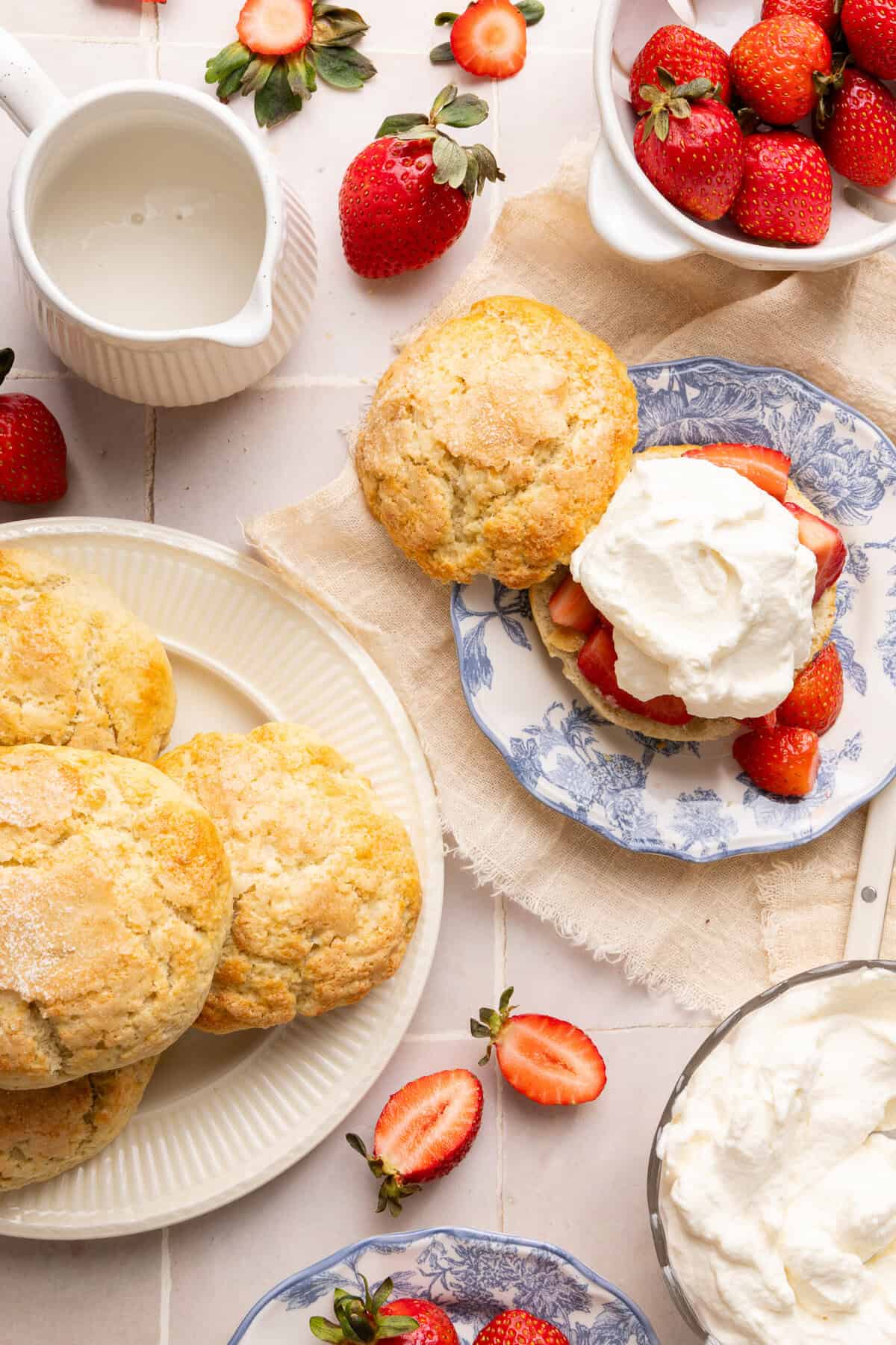 strawberry shortcake on a plate surrounded by biscuits, whipped cream, and strawberries in bowls
