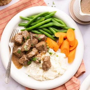 beef tips over mashed potatoes with green beans and carrots on a plate