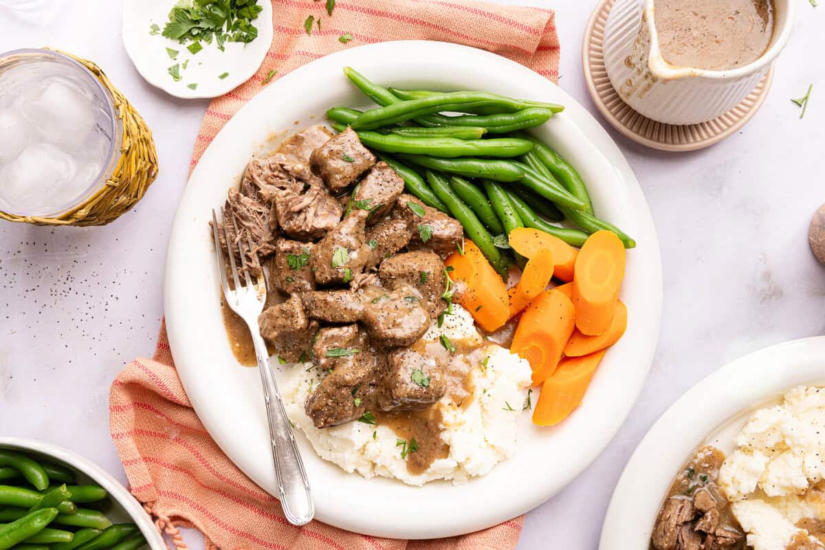 a plate of beef tips over mashed potatoes with vegetables