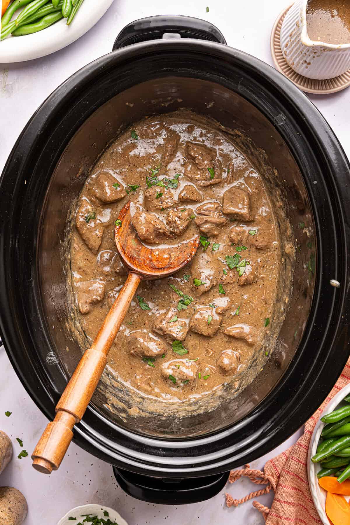 slow cooker beef tips with gravy topped with parsley in a crock pot with a wooden spoon