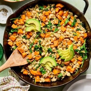 an egg-free breakfast hash with sweet potatoes, ground turkey, and spinach in a cast iron skillet topped with avocado with a wooden spoon