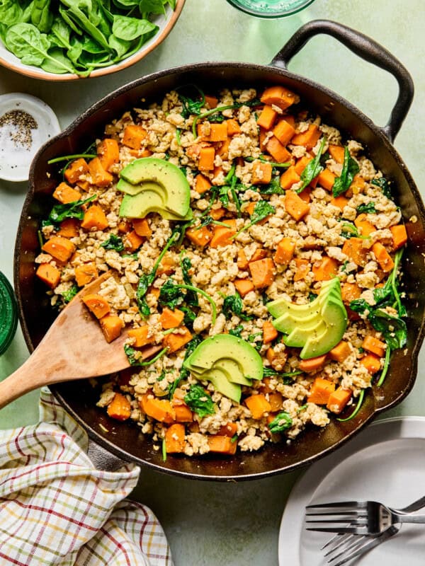 an egg-free breakfast hash with sweet potatoes, ground turkey, and spinach in a cast iron skillet topped with avocado with a wooden spoon