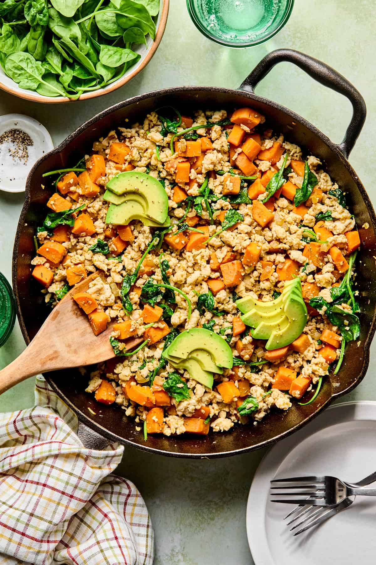 an egg-free breakfast hash with sweet potatoes, ground turkey, and spinach in a cast iron skillet topped with avocado with a wooden spoon