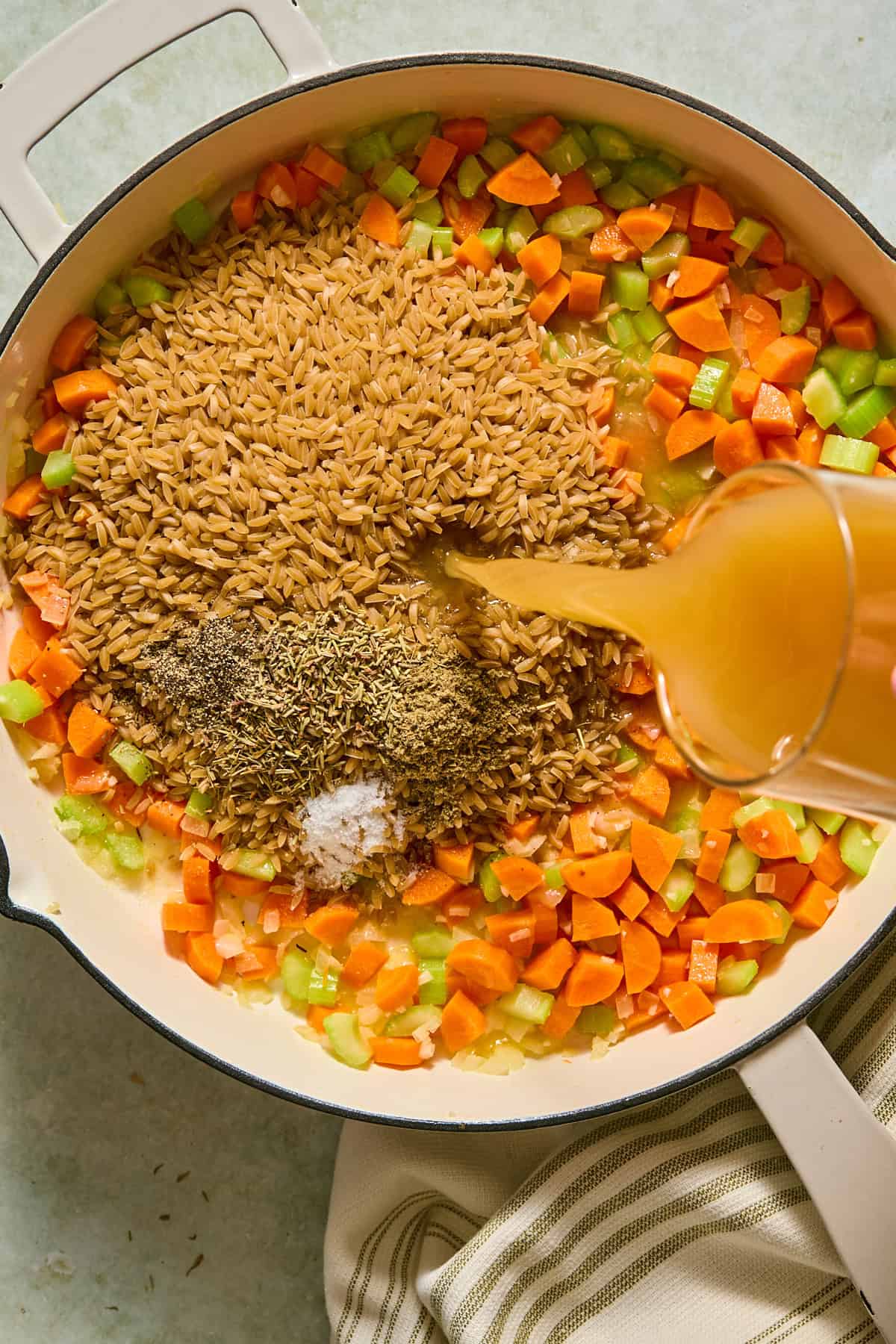 Orzo, sage, thyme, rosemary, salt, and pepper added to a large skillet with softened vegetables as broth is poured in.
