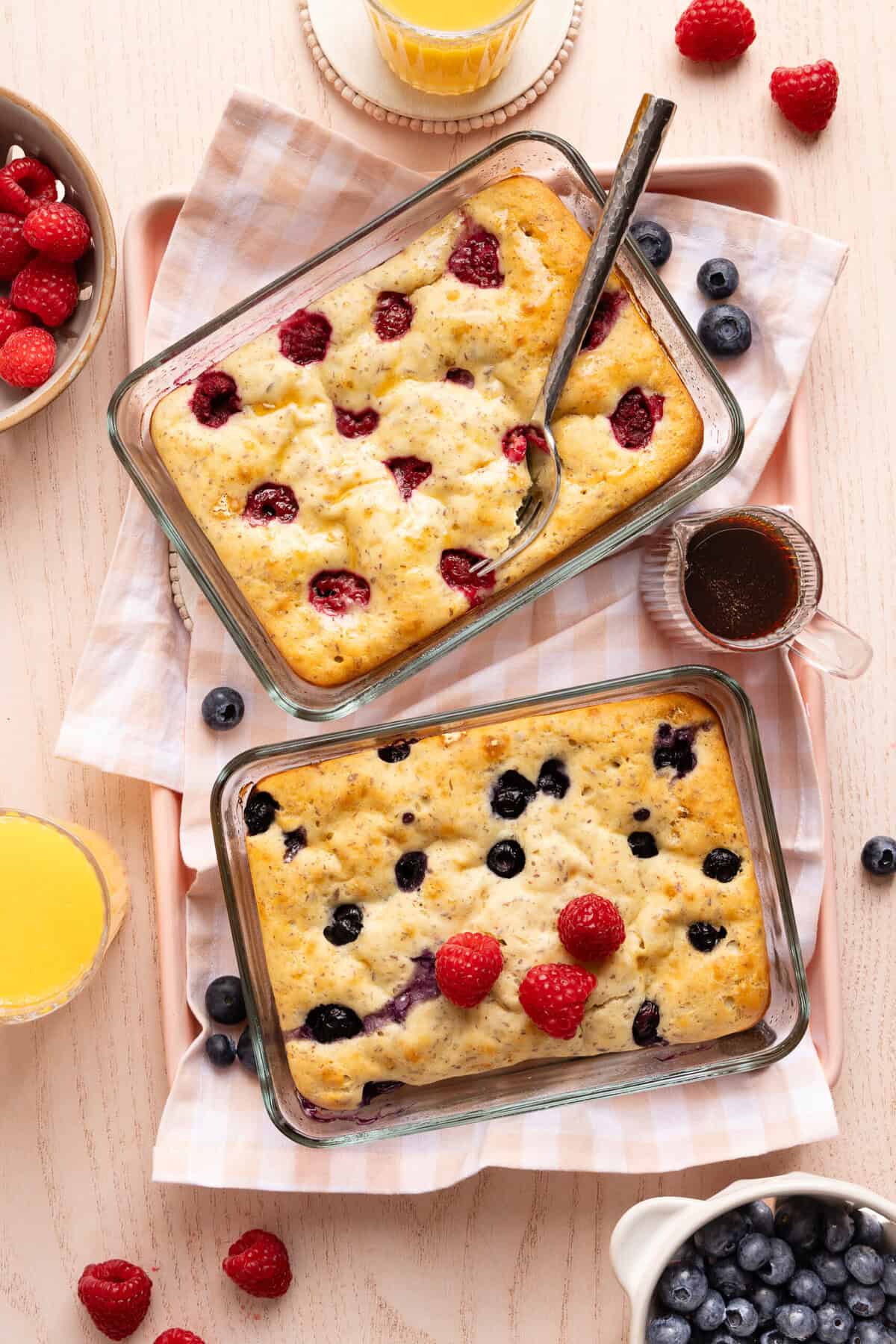 Two Pyrex glass containers filled with baked protein pancake bowls, one topped with raspberries and the other with blueberries. A fork is digging into the raspberry bowl, with a small syrup jar between the containers.
