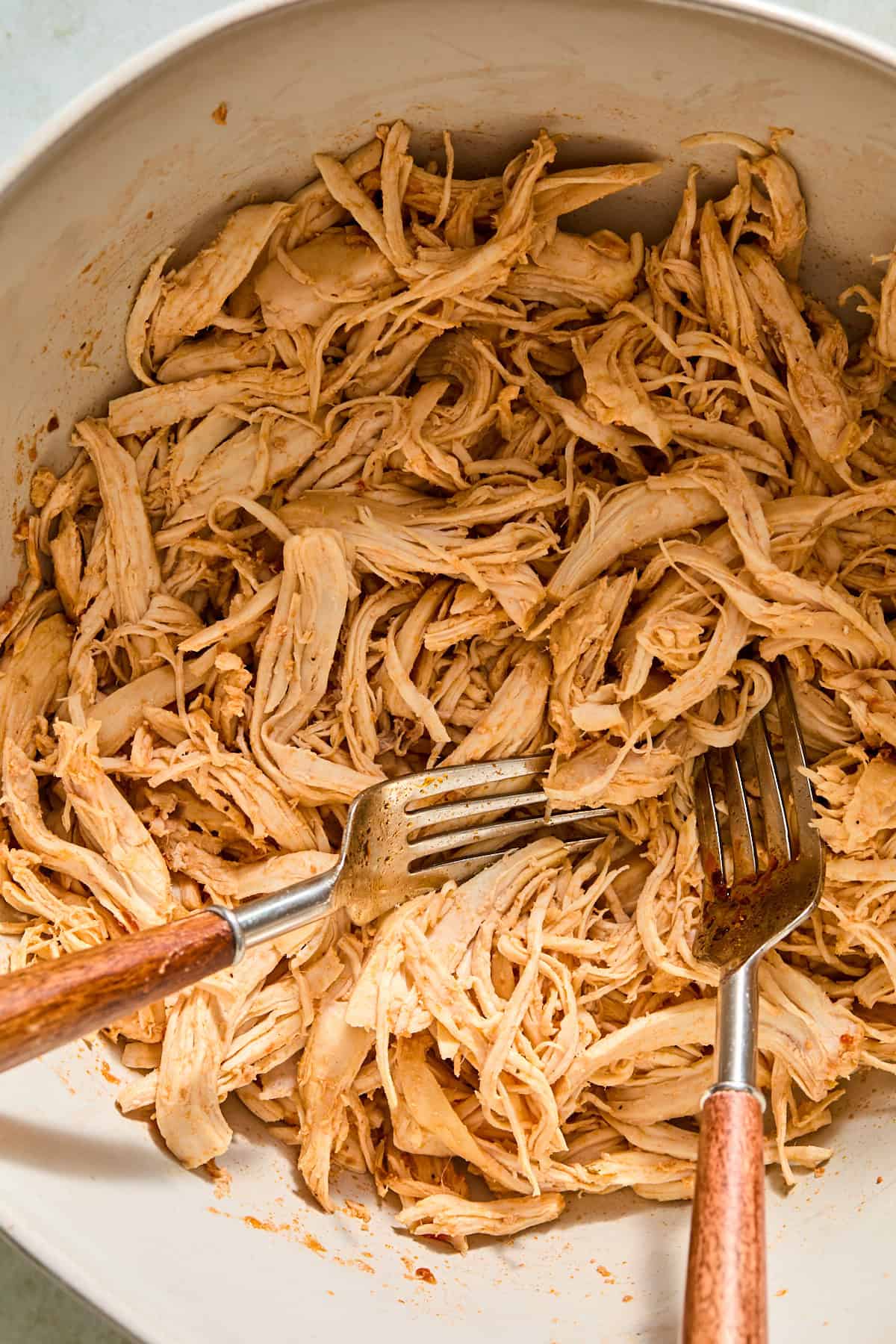 Cooked chicken breasts being shredded with two forks in a mixing bowl.