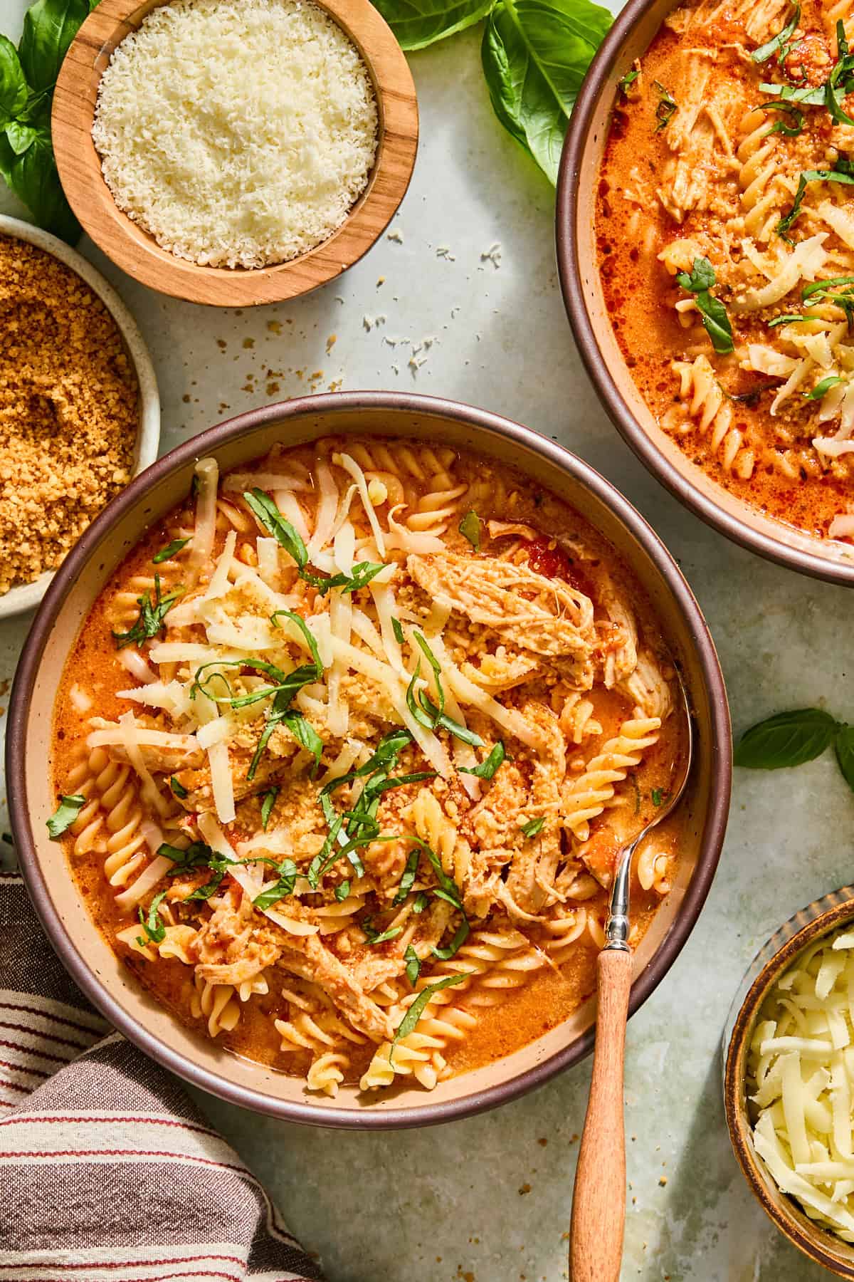 Overhead shot of a bowl of slow cooker chicken parmesan soup with shredded chicken and fusilli, topped with toasted panko, mozzarella, and fresh basil