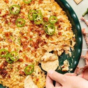 high protein jalapeno popper dip in a green casserole dish with a hand scooping some up with a chip
