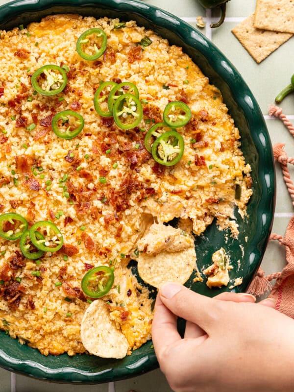 high protein jalapeno popper dip in a green casserole dish with a hand scooping some up with a chip