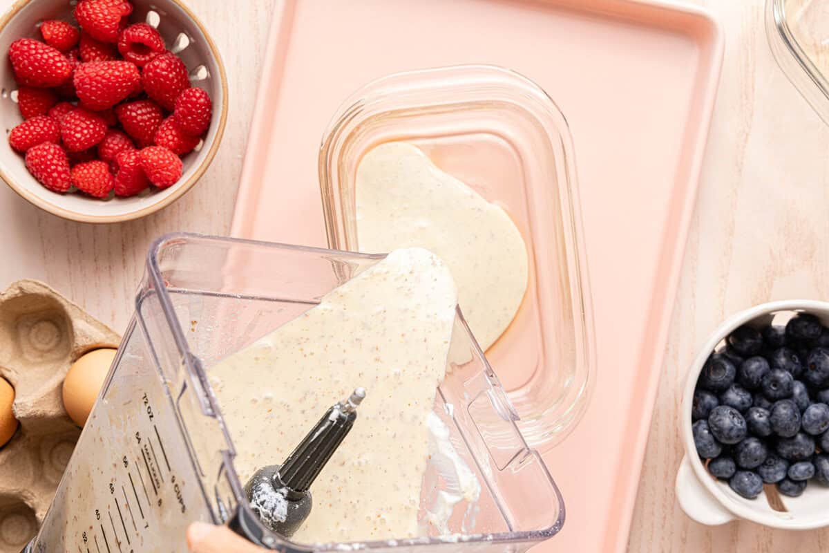 Blender pouring protein pancake batter into a Pyrex glass container, with small bowls of raspberries and blueberries nearby.