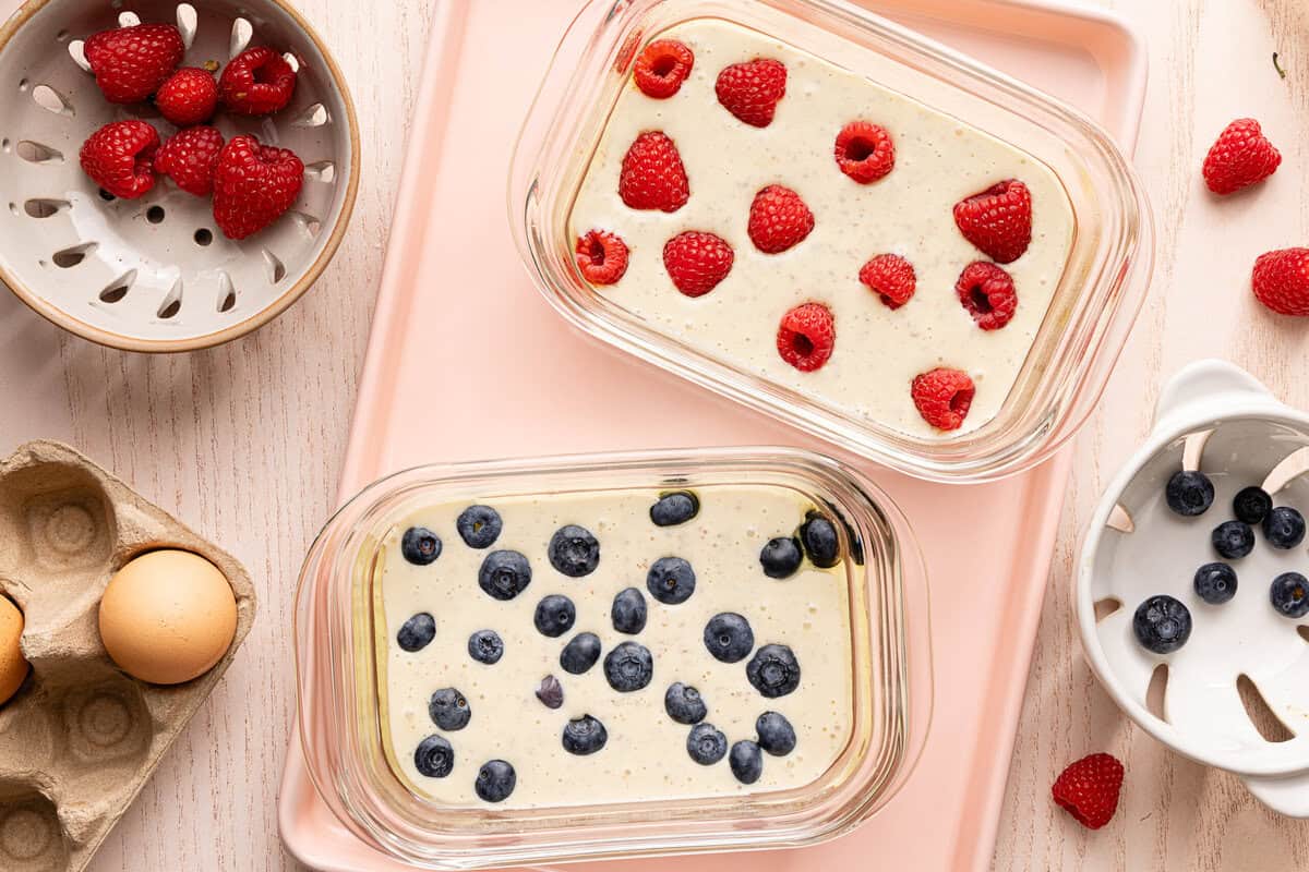 Two Pyrex glass containers filled with protein pancake batter on a countertop, one topped with raspberries and the other with blueberries.