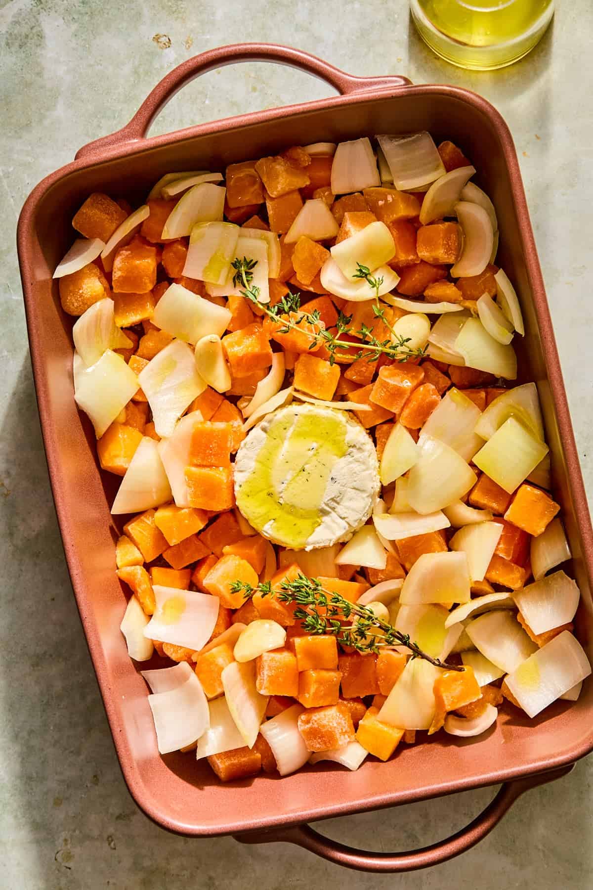 A baking dish filled with frozen butternut squash, veggies, fresh herbs, and a wheel of boursin cheese drizzled with olive oil.