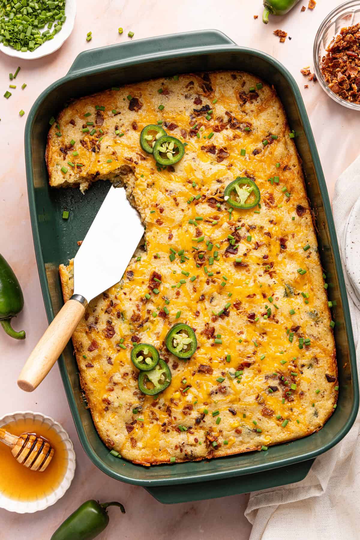 Gluten free cornbread casserole baked in a green 9x13 dish with a slice taken out, topped with melted cheddar, bacon, chives, and jalapeno slices