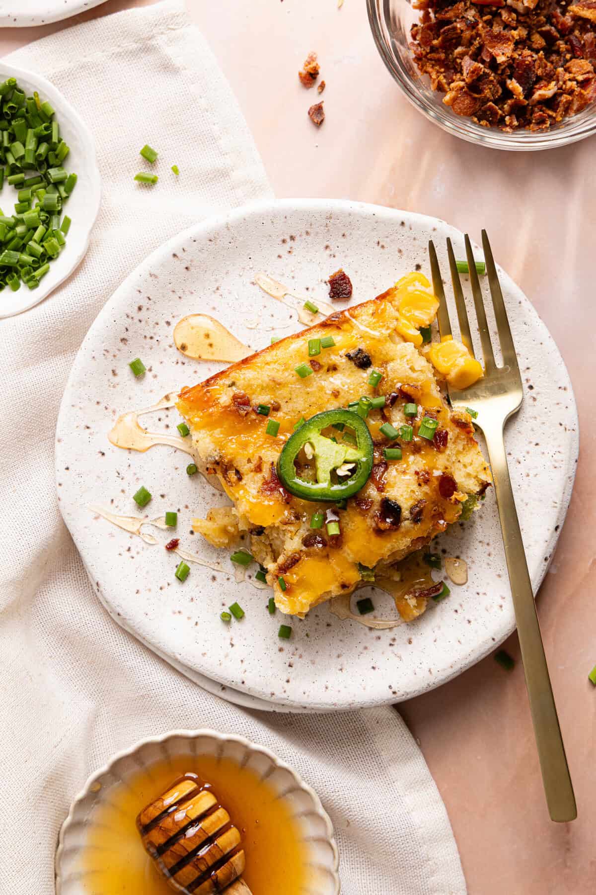 One square of cornbread casserole on a plate with hot honey drizzled over the top and a jalapeño resting in the middle.